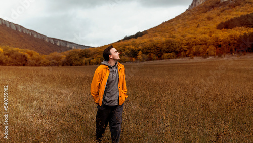Young smiling millennial man in a yellow jacket walks near the picturesque mountains and autumn forest in October