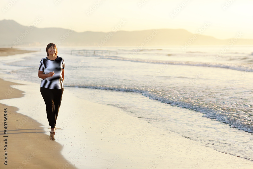 Smiling middle aged woman running on the beach on sunrise. 40s or 50s attractive mature lady in sports clothes doing jogging workout enjoying fitness and healthy lifestyle at beautiful sea landscape.