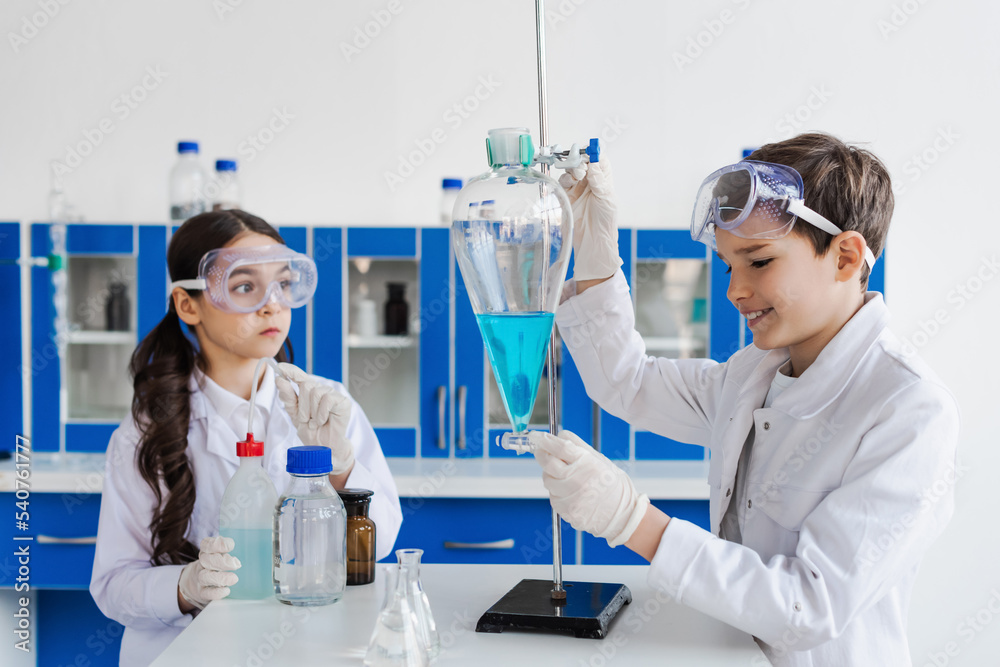 smiling boy holding flask with liquid near serious girl in goggles. smiling boy holding flask with liquid near serious girl in goggles.