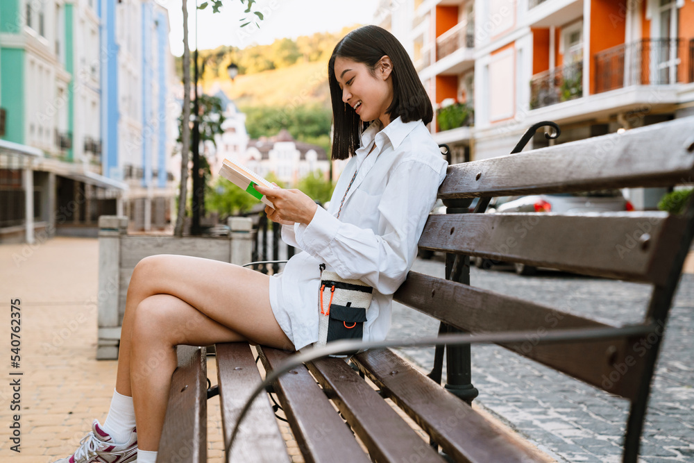 Brunette asian woman smiling and reading book while sitting on bench Brunette asian woman smiling and reading book while sitting on bench