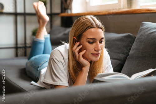 Happy white middle-aged woman reading book while lying on sofa at home