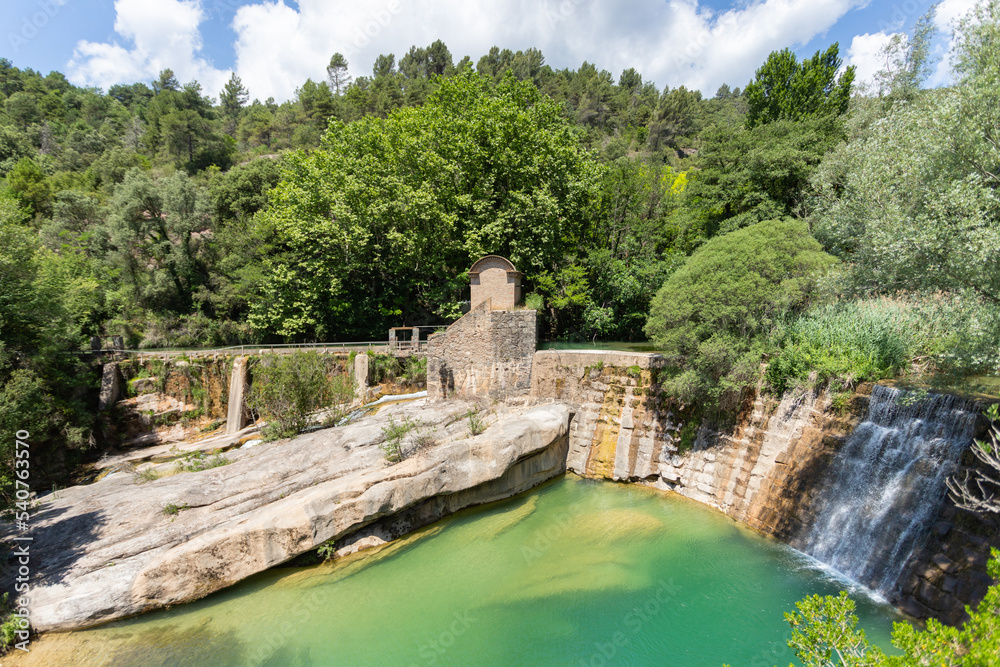Half-height view of the old power plant by a waterfall in a natural park that is transformed into a pond. The river falls like a waterfall into a green pond from a stone wall full of vegetation 