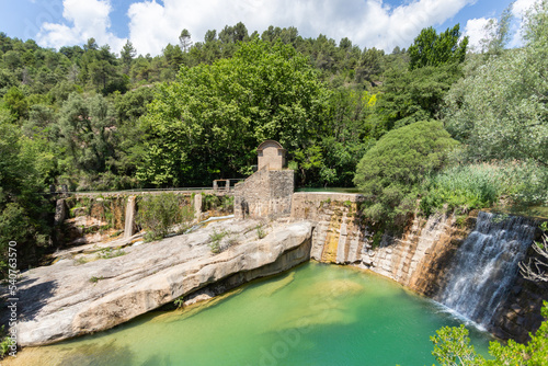 Half-height view of the old power plant by a waterfall in a natural park that is transformed into a pond. The river falls like a waterfall into a green pond from a stone wall full of vegetation 