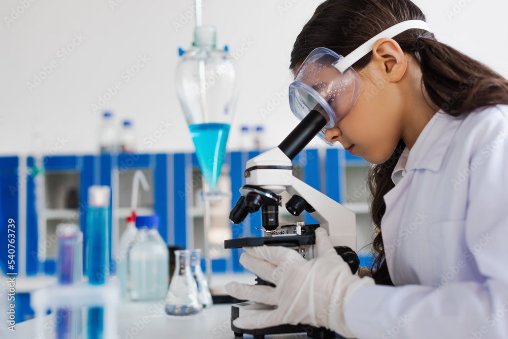 Fototapeta premium side view of preteen girl in goggles looking into microscope in chemical laboratory.