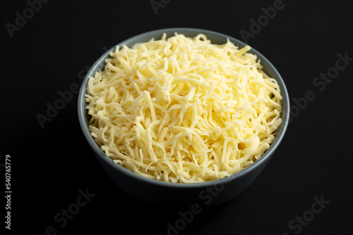 Shredded Mozzarella Cheese in a Bowl on a black surface, side view.