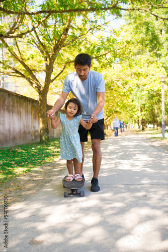 Portrait of Asian father and his daughter skateboarding in park. Happy man holding little girls hands walking on alley while she standing on skateboard. Active rest with kids and fatherhood concept