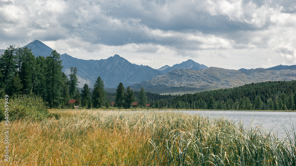 Fototapeta premium Atmospheric view of a mountain lake with camping on the shore in a coniferous forest against the backdrop of mountains and a dramatic cloudy sky