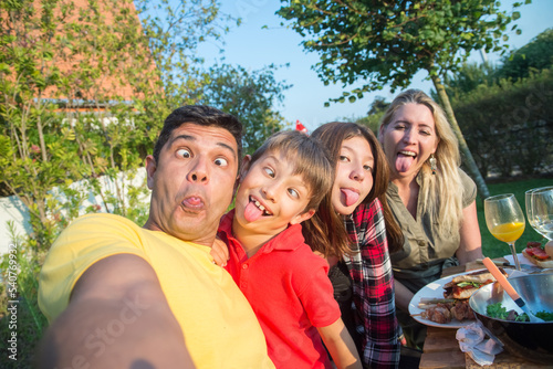 Funny family taking selfie at BBQ party. Mid adult parents and daughter sitting around table, making faces, laughing. BBQ, social media, food, family concept