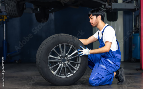 Asian handsome male mechanic wearing uniform, changing rubber tyre or tire wheel, working, reparing in garage at car or automobile maintenance service center or shop with copy space. Industry Concept.