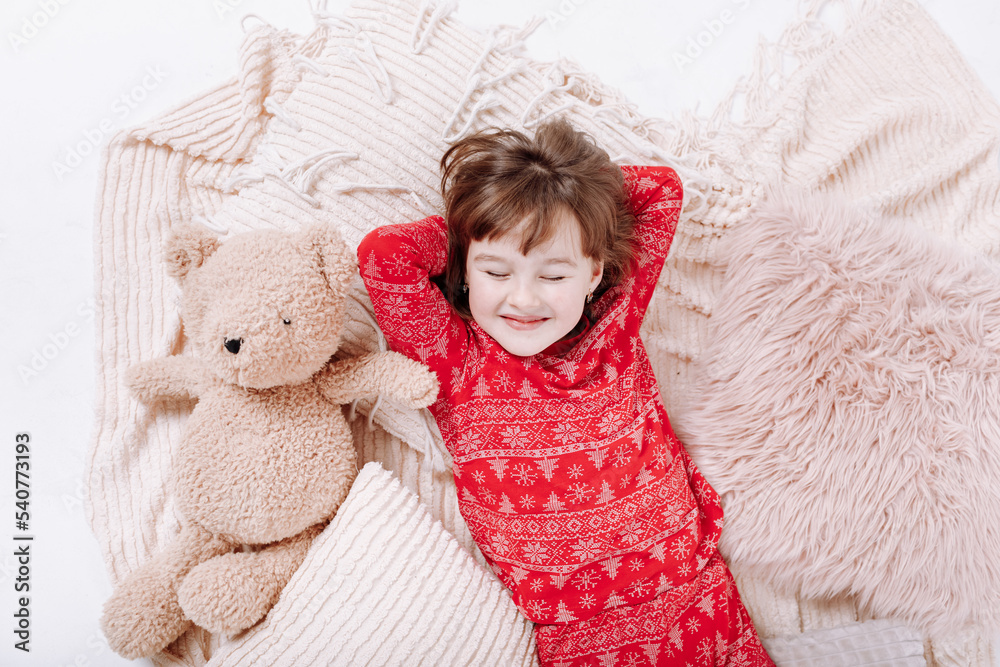 Little smiling girl with closed eyes in red Christmas pyjamas with a Teddy bear are lying on the floor on soft plaid and dreaming about gifts at home. Happy Christmas and new year family holiday.