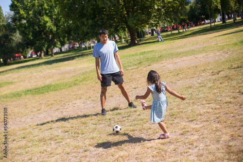 Portrait of Asian father and his daughter playing ball. Handsome man in casual clothes standing on field in park looking at little girl kicking ball. Parenthood and kids healthy growing-up concept ..