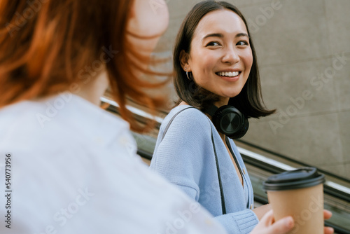 Young multinational women smiling and talking on escalator with coffee