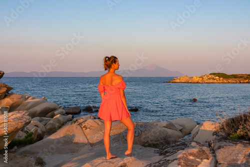 Woman in red dress standing on rock during sunset with scenic view of Mount Athos seen from Karydi beach, Sithonia, Chalkidiki (Halkidiki), Greece, Europe. Summer vacation at Aegean Mediterranean Sea photo