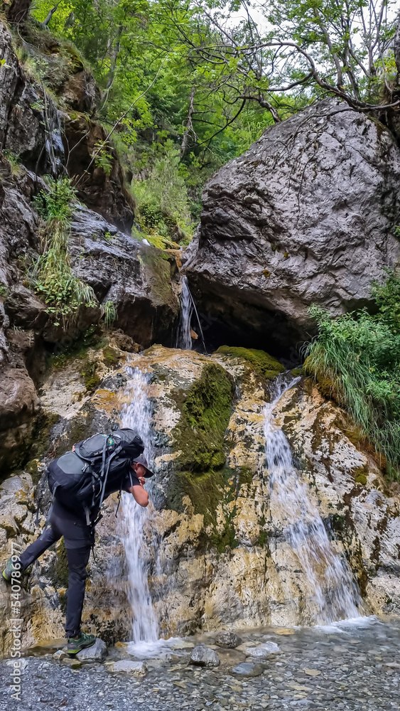 Man with hiking backpack drinking water from famous waterfall located in the Olympus mountains, where God Zeus used to bath. Waterfall at Mount Olympus, Thessaly, Greece, Europe. Shower of Zeus