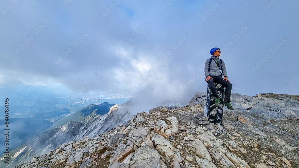 Man with helmet sitting on cloud covered mountain summit of Skolio peak on Mount Olympus, Mt Olympus National Park, Macedonia, Greece, Europe. View of rocky ridges and highlands from throne of Zeus