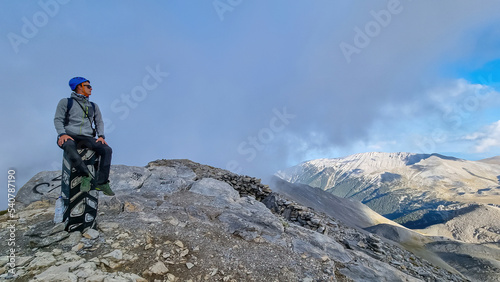 Man with helmet sitting on cloud covered mountain summit of Skolio peak on Mount Olympus, Mt Olympus National Park, Macedonia, Greece, Europe. View of rocky ridges and highlands from throne of Zeus