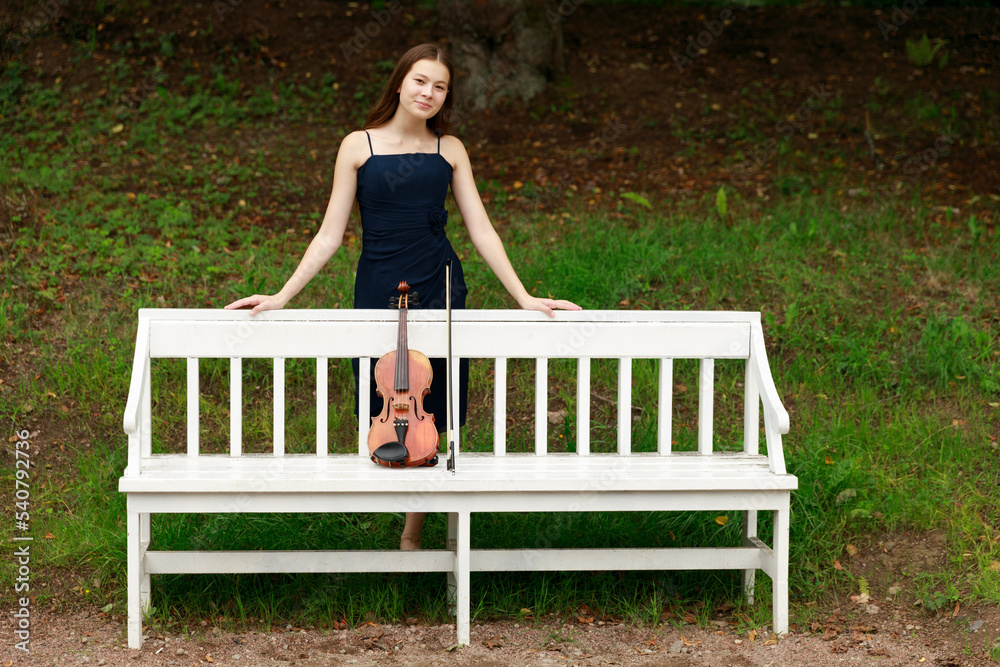 a girl with a violin stands near a white park bench a girl with a violin stands near a white park bench