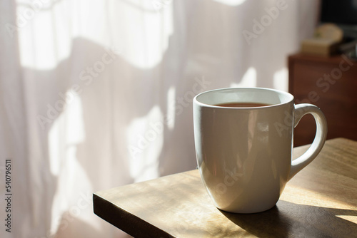 A mug full of coffee or tea set on a wooden table, morning sun and in the background light pink curtains
