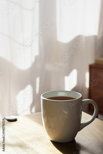 A mug full of coffee or tea set on a wooden table, morning sun and in the background light pink curtains