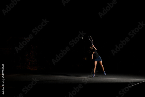 view of female tennis player with racket playing tennis and hitting ball on tennis court at dark time.