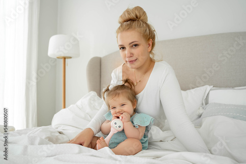 Laughing Mom and baby girl are lying in bed, enjoying time together. Family time concept. Funny playing with toy. Mock up, copy space. Positive babyhood and parenthood.