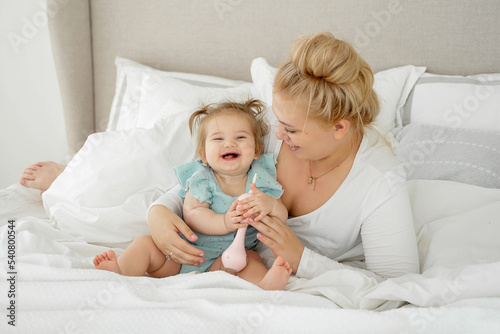 Laughing Mom and baby girl are lying in bed, enjoying time together. Family time concept. Funny playing with toy.