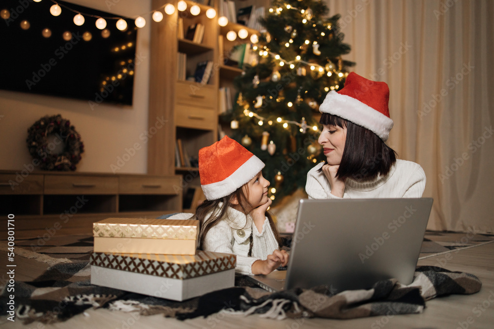 Indoor shot of beautiful happy young woman and her girl shopping online on laptop in cozy Christmas interior. Mother on the floor next the Christmas tree and daughter lying near her, shopping gifts. Indoor shot of beautiful happy young woman and her girl shopping online on laptop in cozy Christmas interior. Mother on the floor next the Christmas tree and daughter lying near her, shopping gifts.