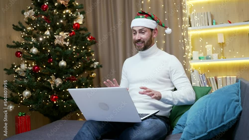 Handsome man in a Santa hat is talking online on video call remotely. Happy New Year Christmas Eve using laptop sitting on the couch at home. waves hand congratulates, greets friends parents Handsome man in a Santa hat is talking online on video call remotely. Happy New Year Christmas Eve using laptop sitting on the couch at home. waves hand congratulates, greets friends parents