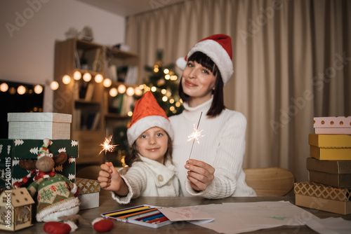 Focus on hands with sparkles, blur background of cheerful cute girl and her mother with charming smile in Santa hats sitting on table on the background of Christmas tree and decorated living room.