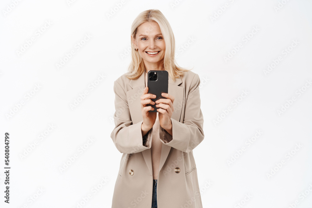 Elegant, beautiful middle aged woman recording video on smartphone, taking photos on mobile phone camera, smiling, standing over white background Elegant, beautiful middle aged woman recording video on smartphone, taking photos on mobile phone camera, smiling, standing over white background