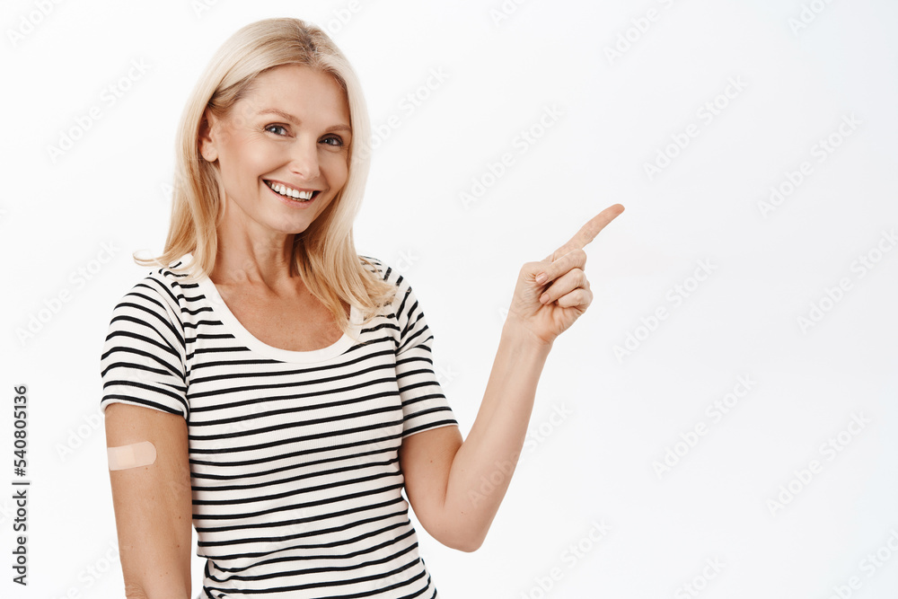 Vaccination among senior people. Smiling mature woman, 50 years old, has band aid from covid-19 vaccination, pointing at banner with coronavirus info, white background Vaccination among senior people. Smiling mature woman, 50 years old, has band aid from covid-19 vaccination, pointing at banner with coronavirus info, white background
