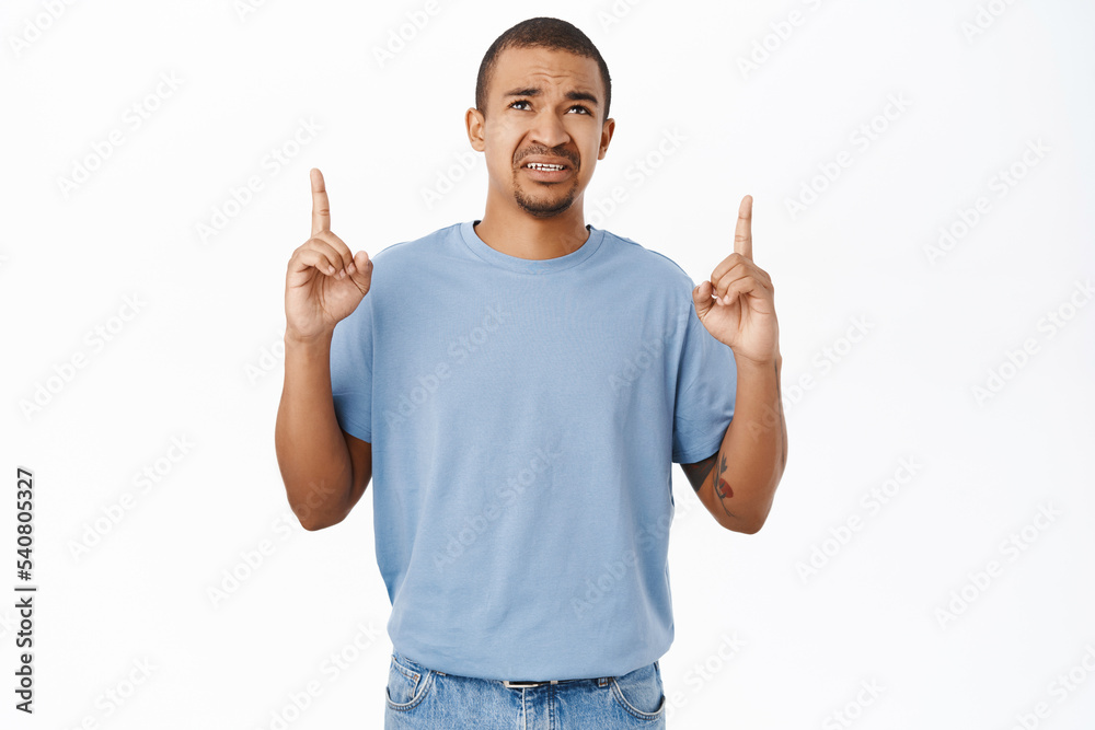 Portrait of young man pointing fingers up, grimacing and looking with dislike, cringe and disappointment, standing over white background
