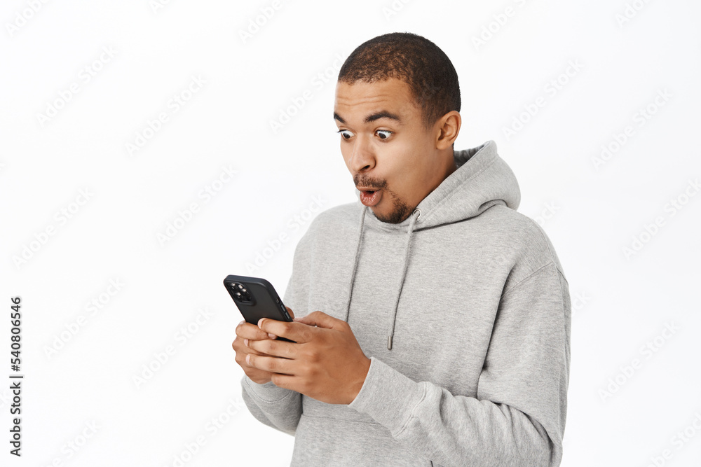 Amazed guy looks at his phone, sees awesome notification on smartphone, stands happy over white background Amazed guy looks at his phone, sees awesome notification on smartphone, stands happy over white background