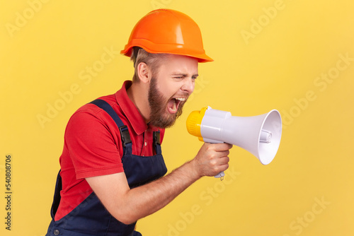Side view of angry bearded worker wearing protective helmet and blue overalls holding megaphone and screaming with aggressive expression, protesting. Indoor studio shot isolated on yellow background.