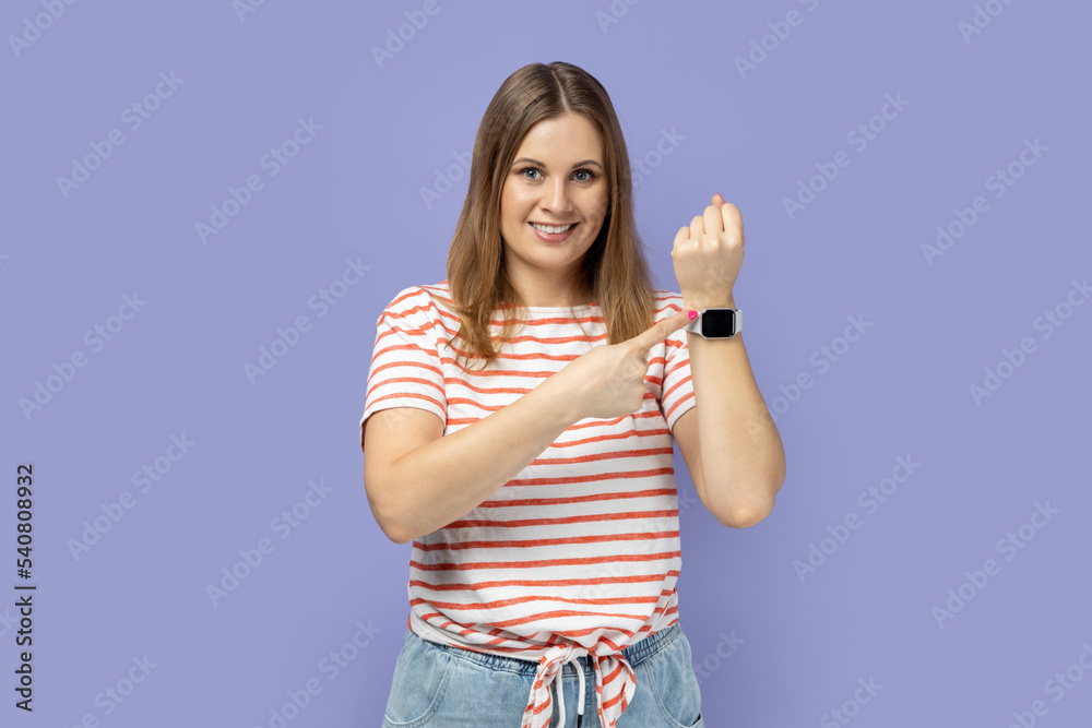 Good result and ontime. Portrait of blond woman wearing striped T-shirt standing, showing his smart watch and looking at camera with toothy smile. Indoor studio shot isolated on purple background. Good result and ontime. Portrait of blond woman wearing striped T-shirt standing, showing his smart watch and looking at camera with toothy smile. Indoor studio shot isolated on purple background.