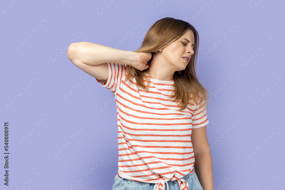 Portrait of attractive tired upset unhealthy blond woman wearing striped T-shirt standing massaging neck to relieve pain, muscle strain in back. Indoor studio shot isolated on purple background. Portrait of attractive tired upset unhealthy blond woman wearing striped T-shirt standing massaging neck to relieve pain, muscle strain in back. Indoor studio shot isolated on purple background.