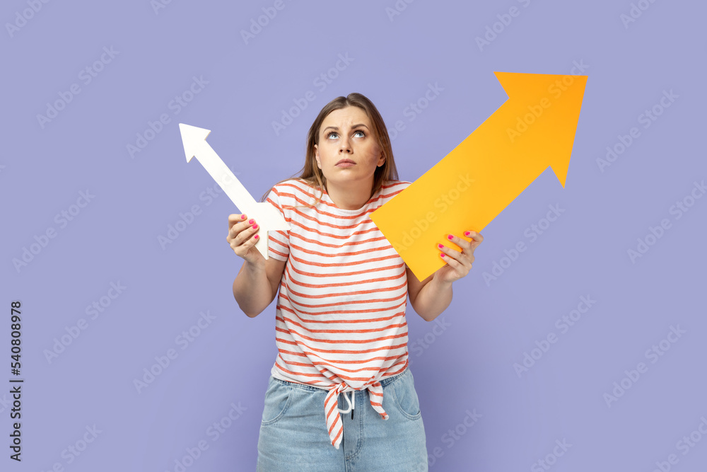 Portrait of confused puzzled blond woman wearing striped T-shirt holding two arrows indicating to different sizes, looking up and shrugging. Indoor studio shot isolated on purple background. Portrait of confused puzzled blond woman wearing striped T-shirt holding two arrows indicating to different sizes, looking up and shrugging. Indoor studio shot isolated on purple background.