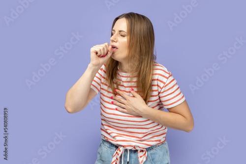 Portrait of sick unhealthy blond woman wearing striped T-shirt having cough and feeling pain in her lungs, catching cold, flu symptoms. Indoor studio shot isolated on purple background.