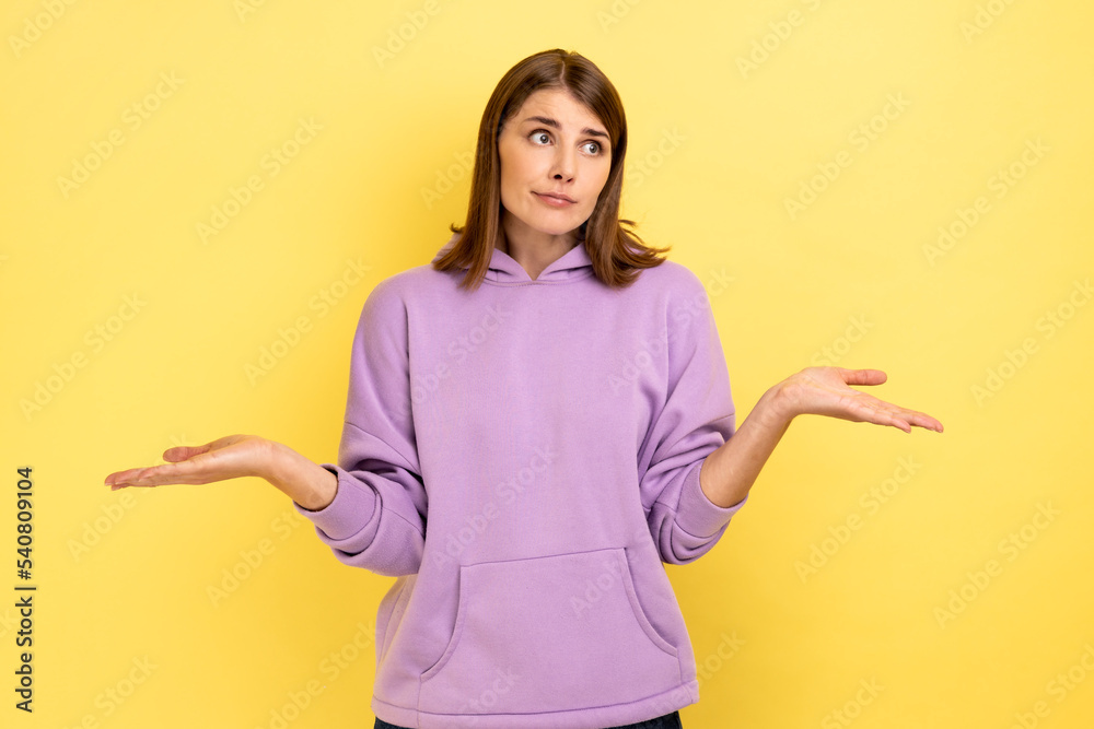 I don't know. Portrait of confused beautiful young woman standing with raised arms and looking at camera, wearing purple hoodie. Indoor studio shot isolated on yellow background.