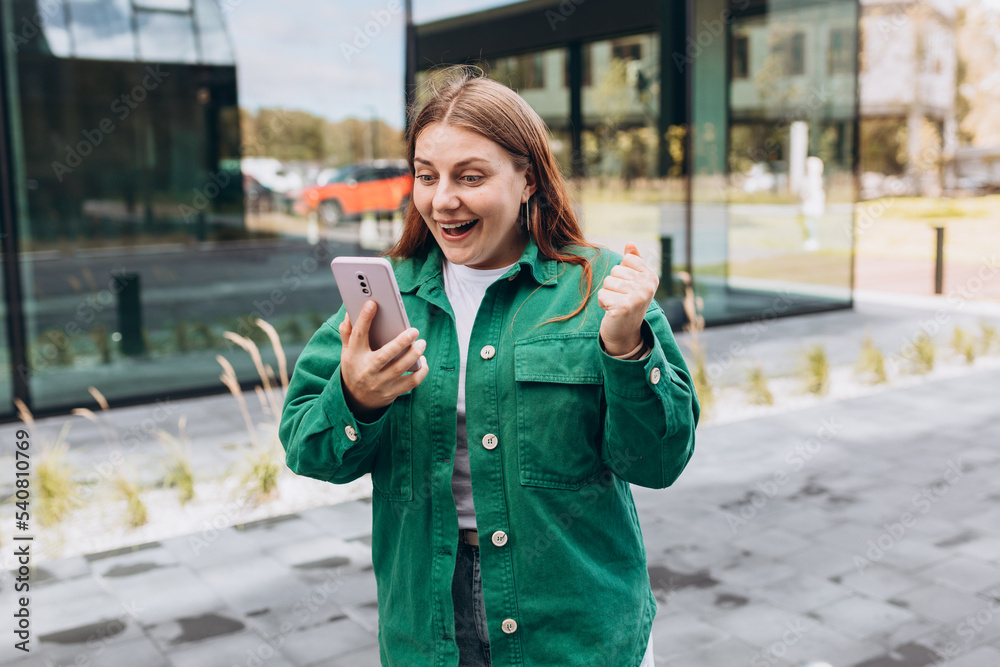 Fototapeta premium Portrait of a satisfied young woman holding mobile phone and celebrating outdoors. Happy girl walking on city street, hipster female overjoyed with winning online contest on website