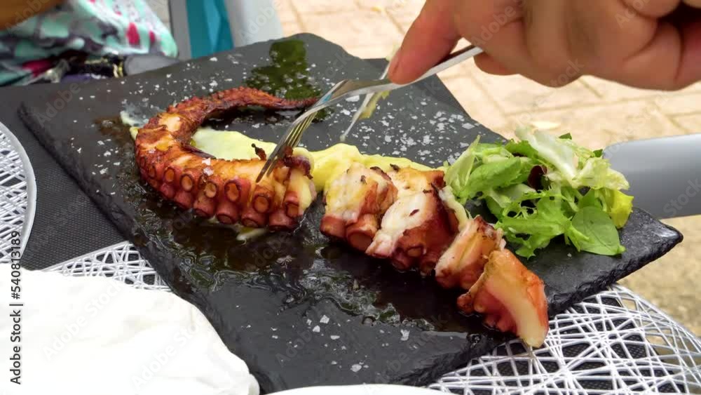 Pata de pulpo sobre plato de pizarra siendo cortada por manos de hombre en una terraza de un restaurante con su molesta mosca cojonera. España