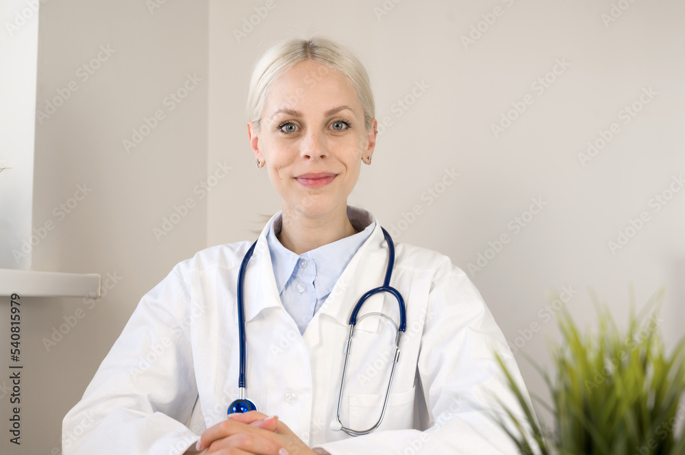 Portrait of smiling young Caucasian female doctor sit at desk in hospital or private clinic in white medical uniform at workplace, healthcare concept Portrait of smiling young Caucasian female doctor sit at desk in hospital or private clinic in white medical uniform at workplace, healthcare concept