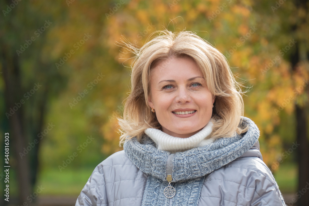 Middle-aged blonde woman on a walk in the autumn park. Portrait of a mature woman smiling at the camera at the park. Psychological health, psychology concept. Middle-aged blonde woman on a walk in the autumn park. Portrait of a mature woman smiling at the camera at the park. Psychological health, psychology concept.