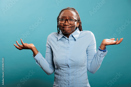 Puzzled and doubtful young adult person being hesitant and unconfident while answering question. Confused and uncertain woman shrugging shoulders with uncertainty while standing on blue background. photo