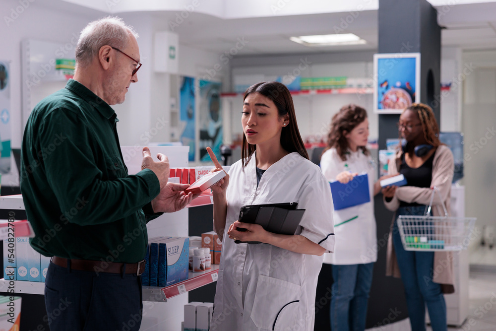 Obraz premium Druggist talking with elderly client during medical consultation in drugstore, explaining pills label. Pharmacist selling drugs to cure cardiology disease. Drugstore shelves full with medication