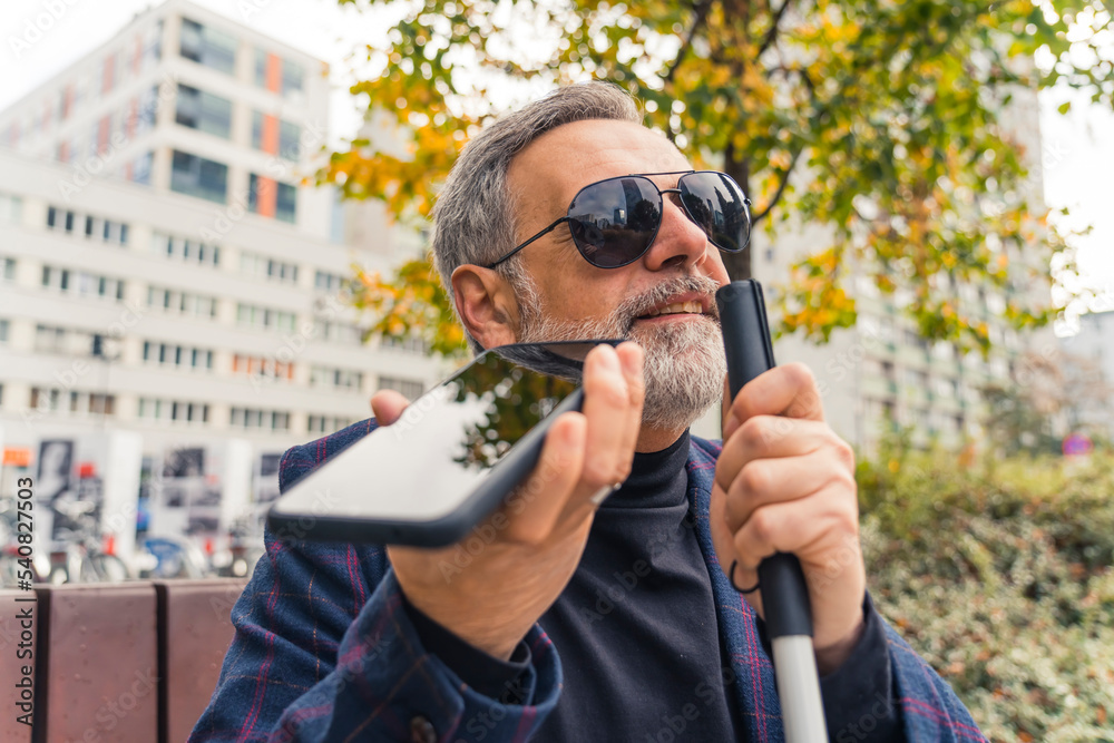 Blind middle-aged man with gray hair and beard wearing sunglasses holding cane and phone using text-to-speech function. Accessibilty. Modern technology. Horizontal outdoor shot. High quality photo - obrazy, fototapety, plakaty 