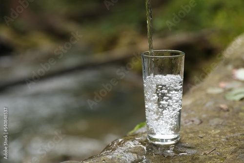 Fresh water pouring into glass on stone near stream. Space for text