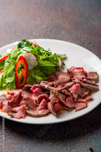 Sliced roast beef with green salad on white plate on dark stone table macro close up vertical