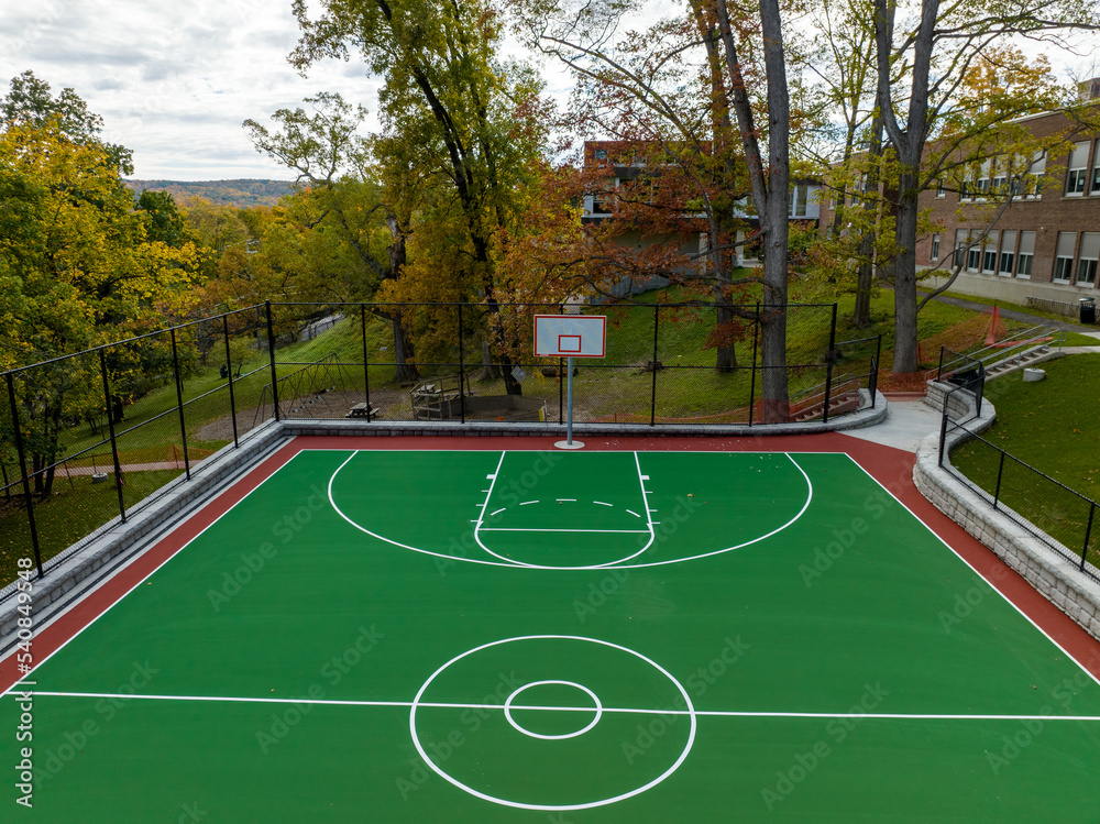 Fototapeta premium Aerial photo of a green and red outdoor basketball court at school playground. Court includes retaining walls and black vinyl coated chain link fence.