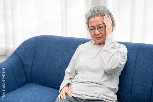 senior woman sitting on a sofa at home with a headache, feeling pain and with an expression of being unwell. Health problem concept.